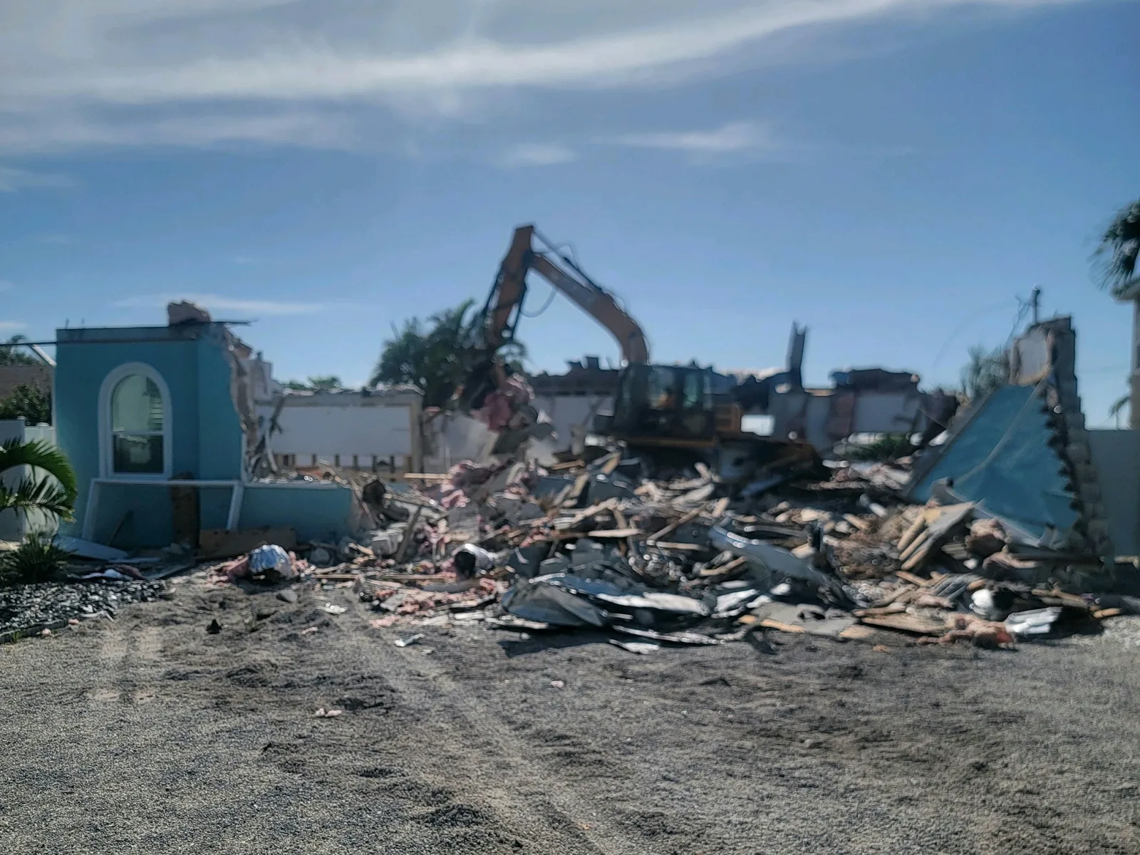 House reduced to a pile of rubble with an excavator working behind it and a blue stucco wall fragment still standing